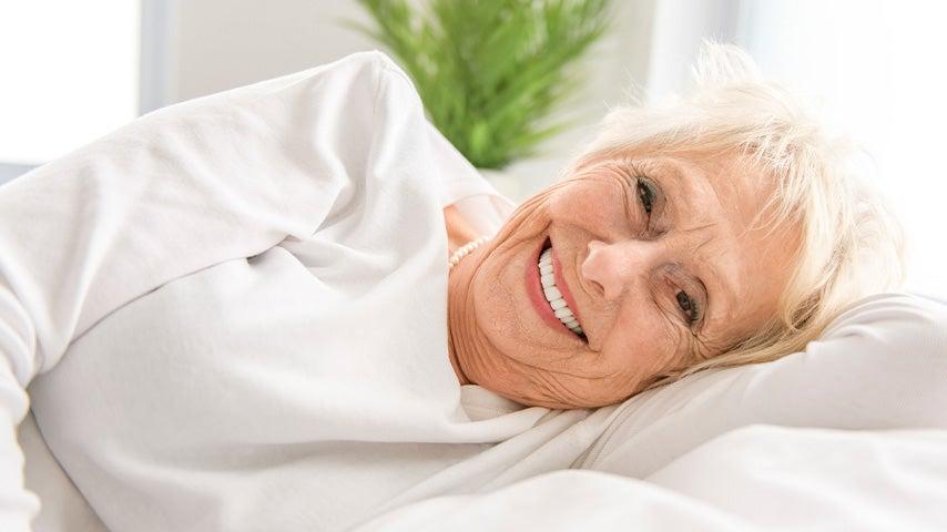 smiling older woman wearing white shirt and pearls lying on bed with white sheets
