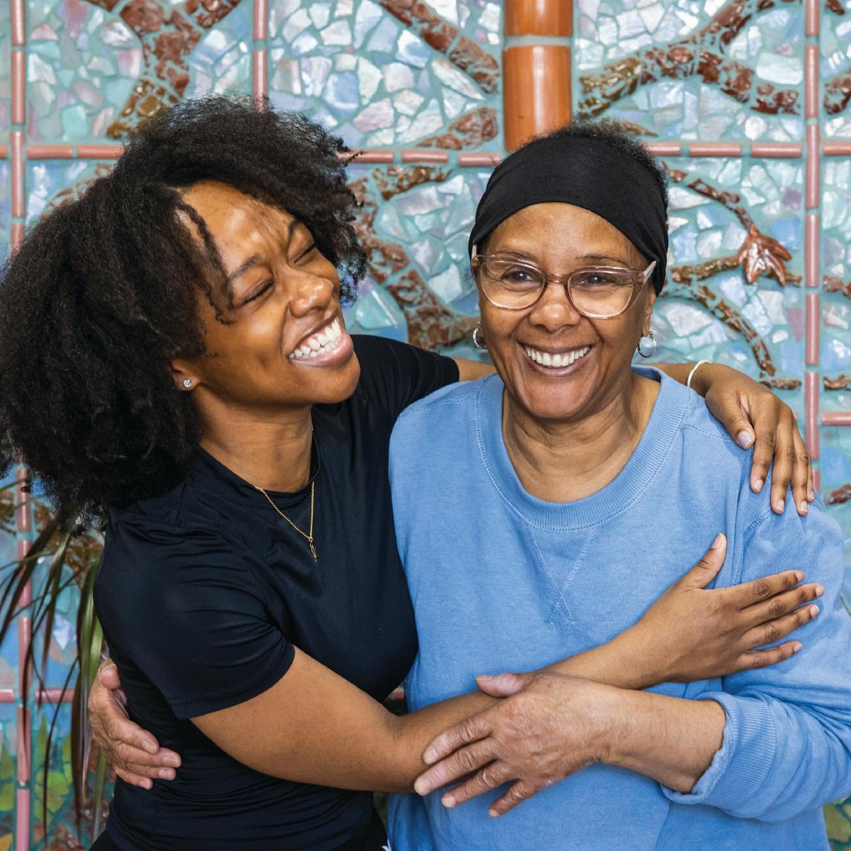 A Black younger woman is embracing her grandmother. Both are laughing/enjoying life.