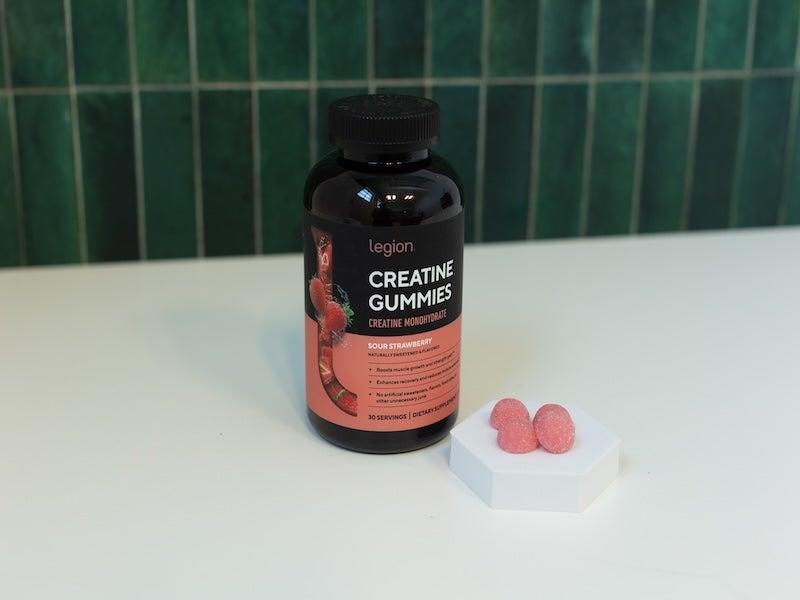 Black bottle of Legion Creatine Gummies, with four pink gumdrop gummies beside it on a white pedestal