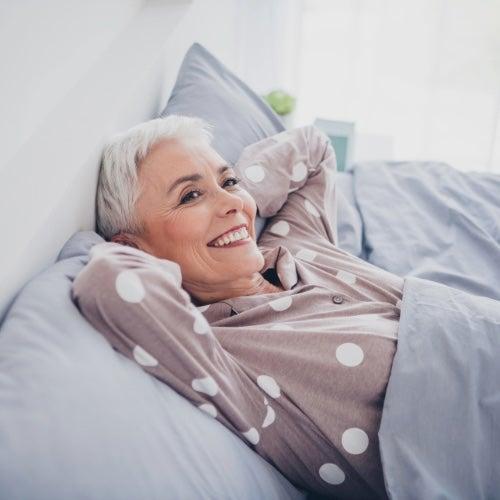 A woman with short white hair and grey polka dot pajamas leans back in bed