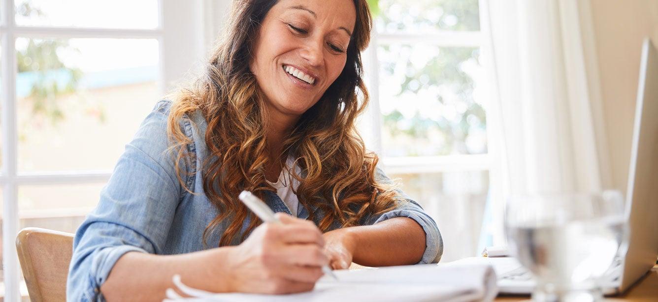 A middle-aged woman is writing in notebook in front of her computer.
