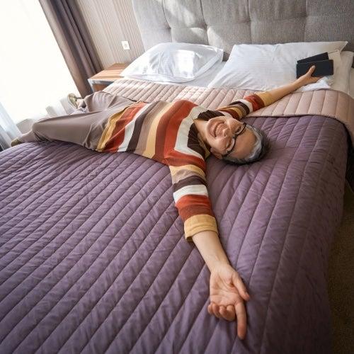 A happy older adult woman spreads out on a mattress with a purple bedspread