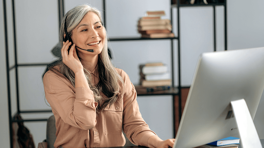 A woman with long gray hair sits in front of a computer with a headset on.
