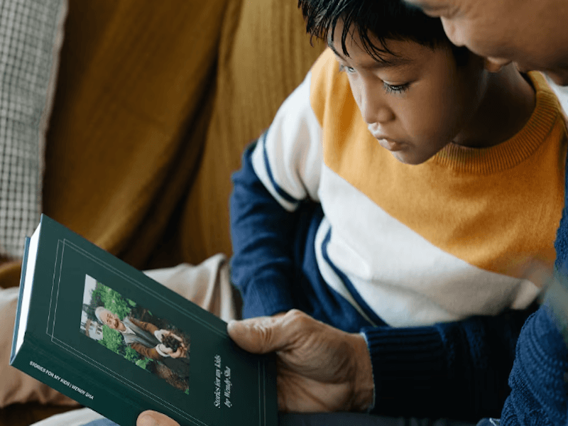 Young boy in a striped sweater looking at a book held by an adult with a photo of an older man on the cover.