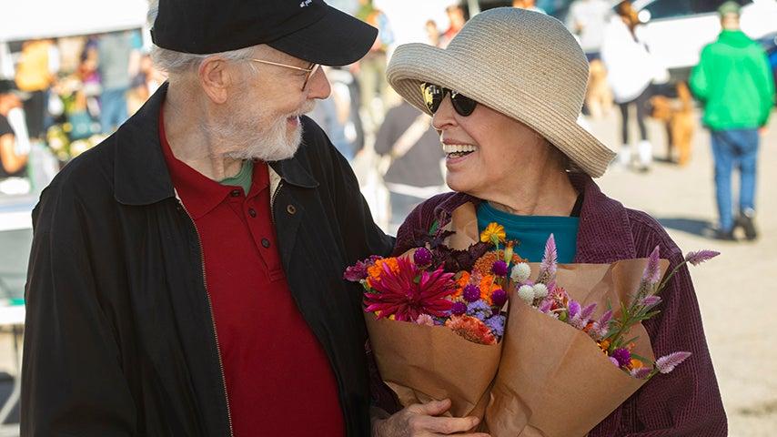 A man and woman smile at each other. The woman is holding two flower bouquets.