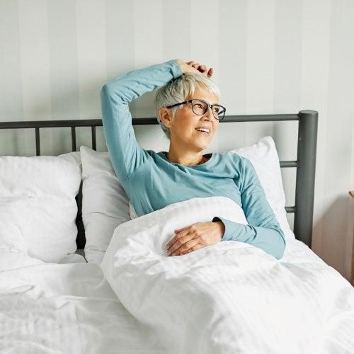A woman in a blue shirt and glasses relaxes in a bed with white sheets