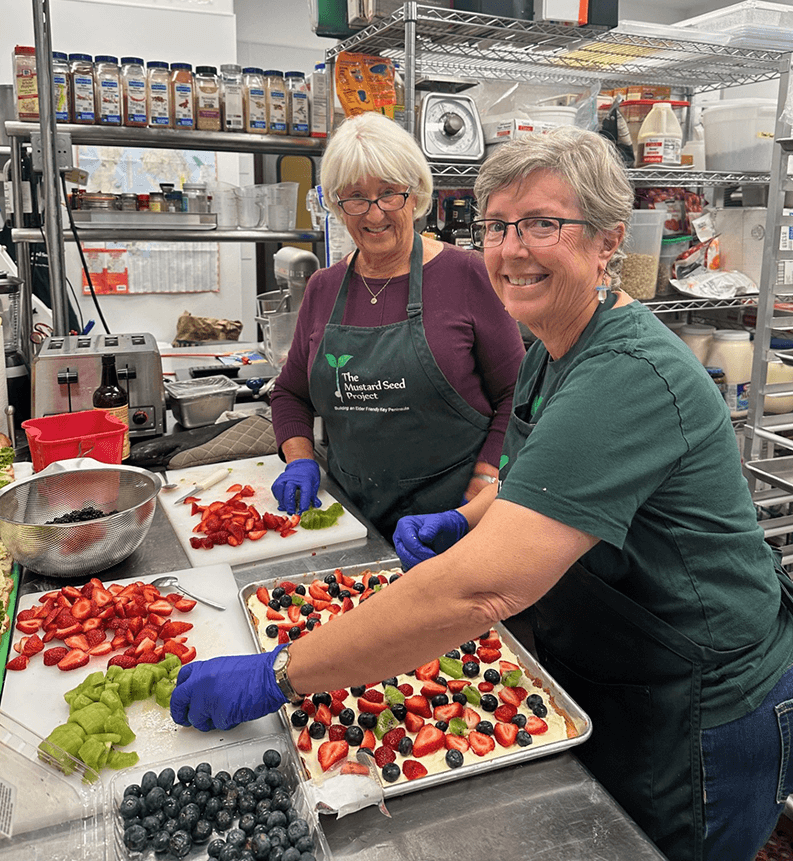 two smiling older women with Mustard Seed Project aprons prepare sliced fruit