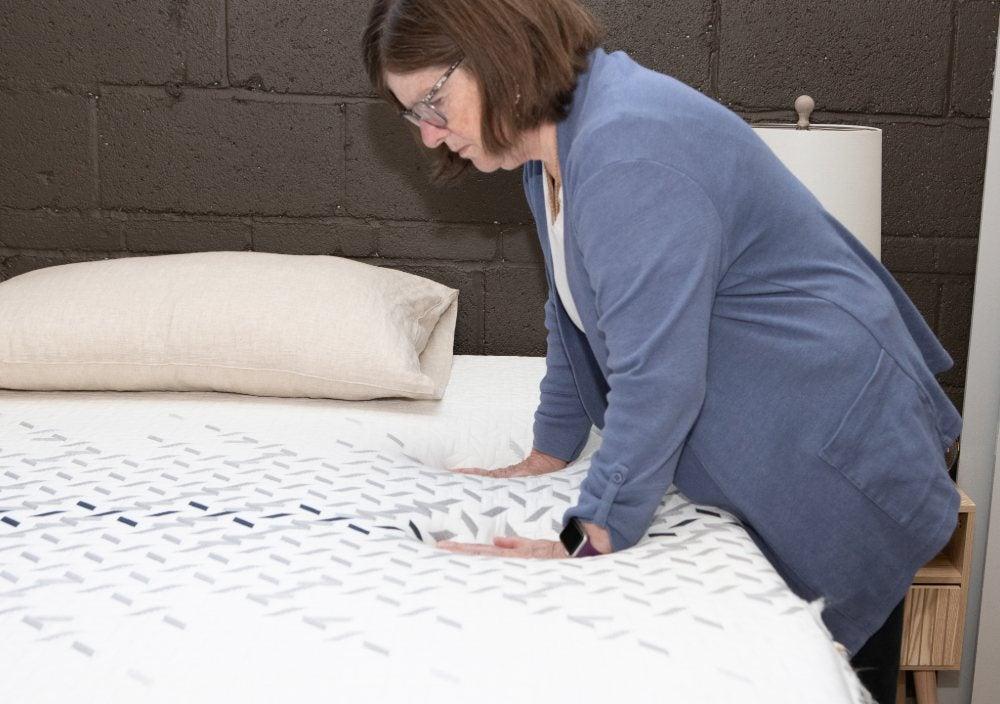 A standing woman presses her hands down onto the surface of a mattress