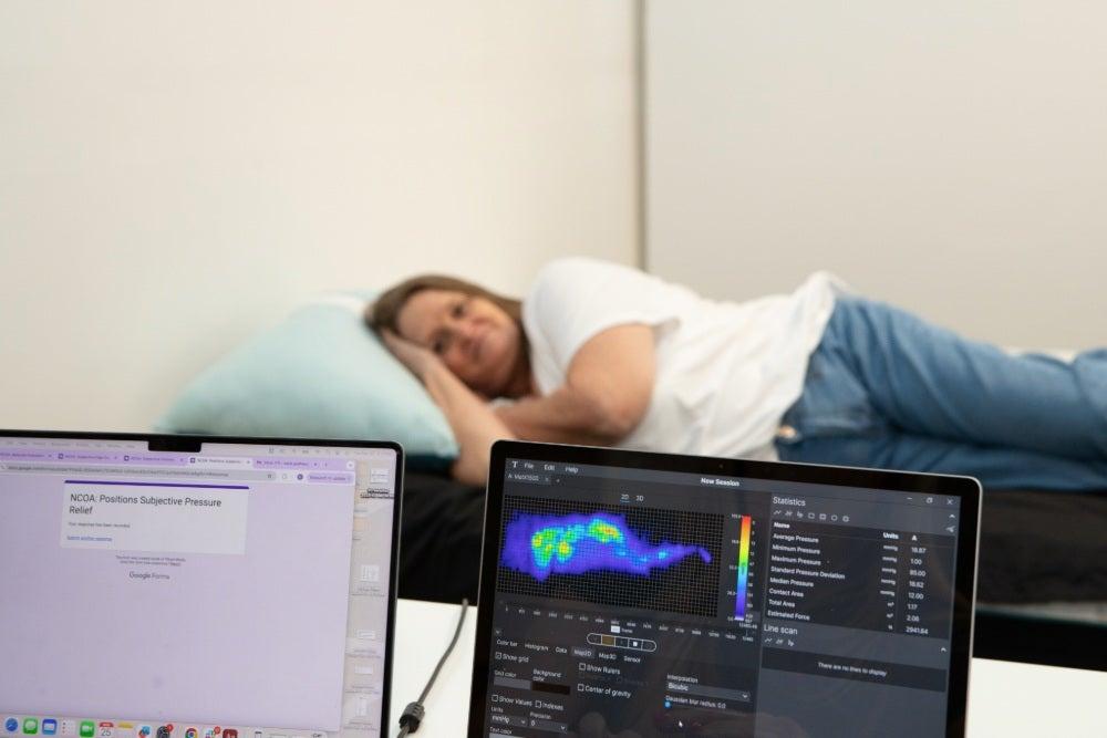An older woman lying on her side on a mattress, with a computer screen showing a pressure map in front of her