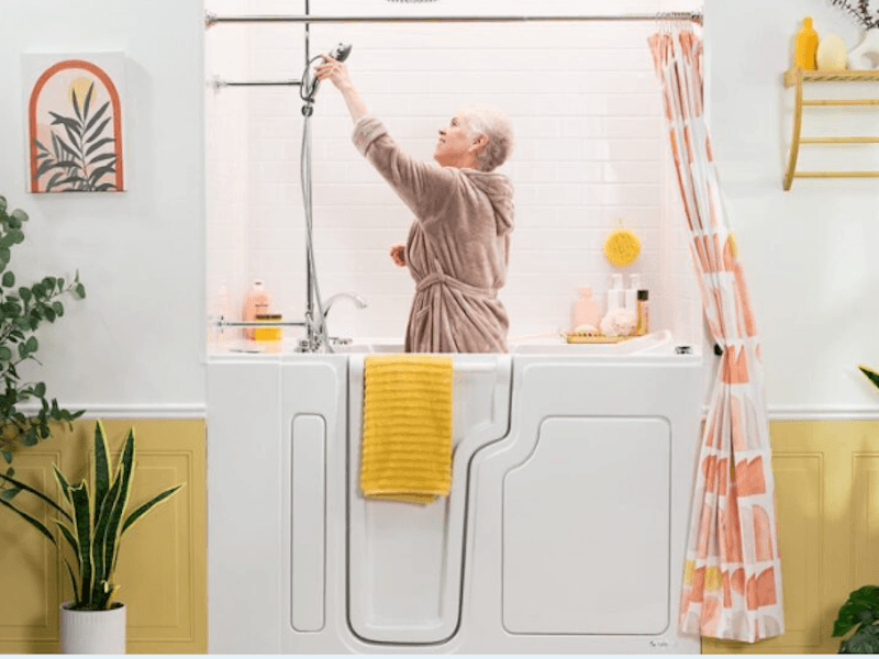 Older woman in a bathrobe adjusts the showerhead in a walk-in bathtub. The setting is cozy with plants, a yellow towel, and patterned shower curtain.