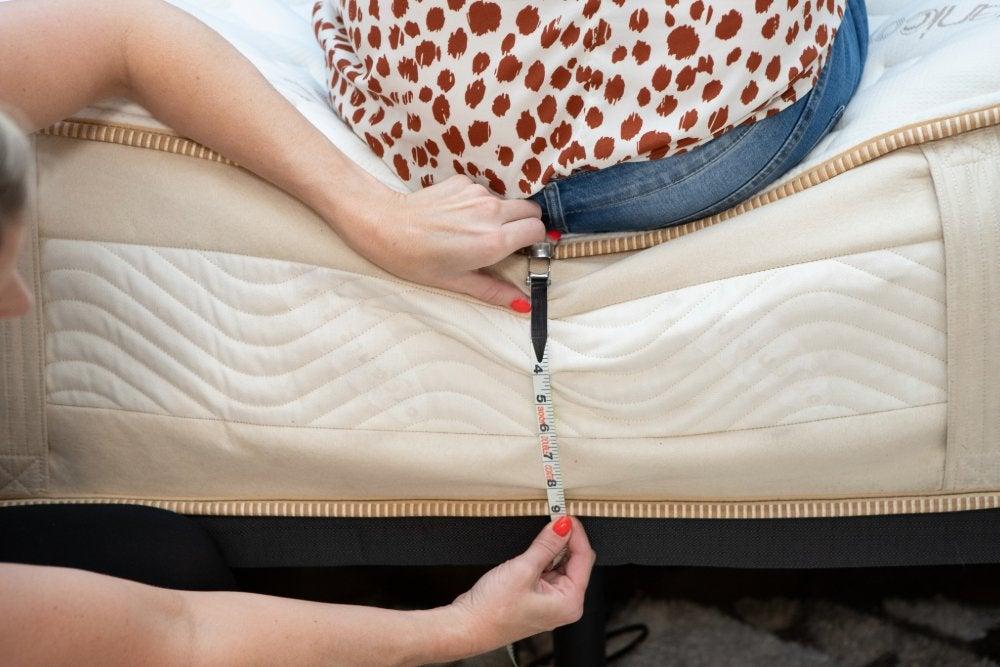 A woman sitting backwards on the edge of a Saatva Rx mattress while another woman measures the edge