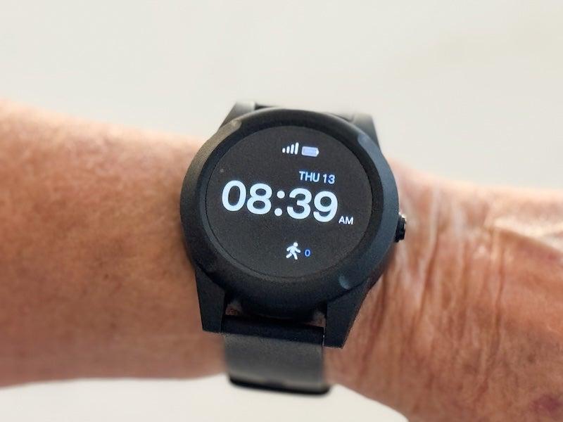 A person displays a black watch on their left wrist; the clock face is slightly larger than the cap of a milk jug, and digitally displays the time 8:39 a.m. in white font, as well as displaying icons for network connection, battery life, and steps.