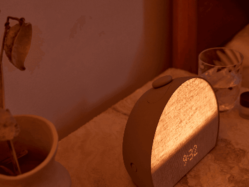 Beside table warmly lit by a dome-shaped alarm clock showing the time ‘9:32.’.