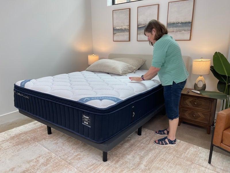 A woman stands to one side of the DreamCloud Premier Hybrid Mattress and presses on it.