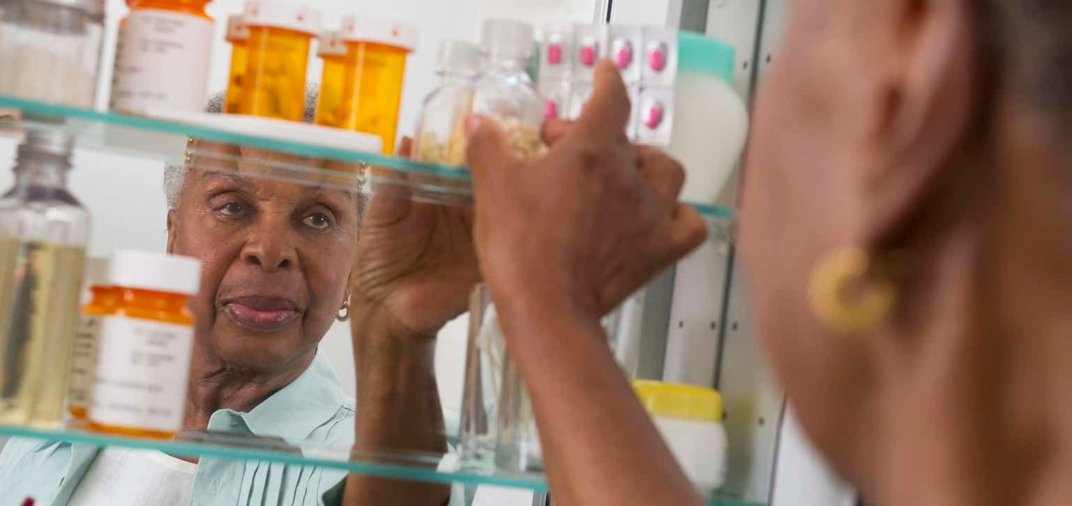 An older black women is reaching for a prescription bottle in her medicine cabinet.