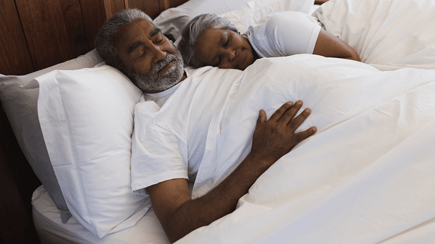 older Indian man and woman sleeping on fluffy white pillows under a white blanket