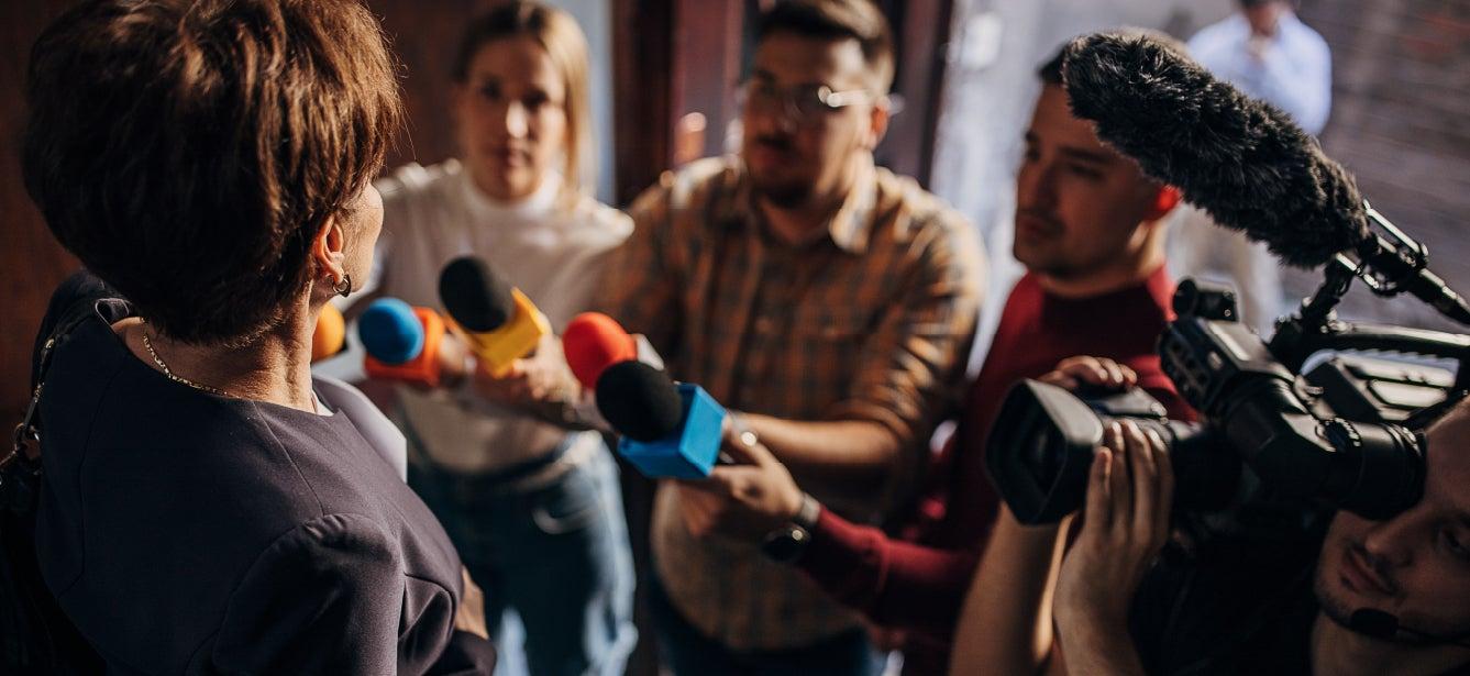 A group of journalists is gathered around a person, likely a public figure, holding out various colorful microphones and a video camera. The setting appears to be indoors with dim lighting. The focus is on the person being interviewed, who is facing away from the camera, while the journalists are captured mid-action, eagerly trying to capture the interview.
