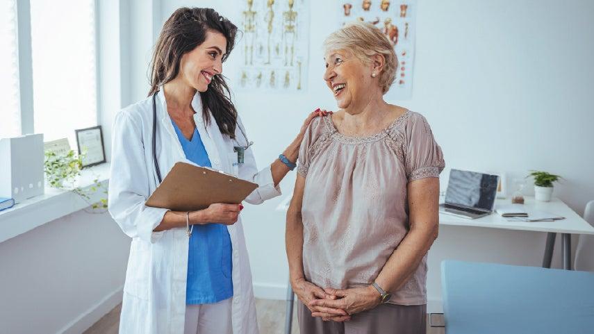 An older caucasian woman is speaking to a younger doctor during her doctor's visit, both are smiling