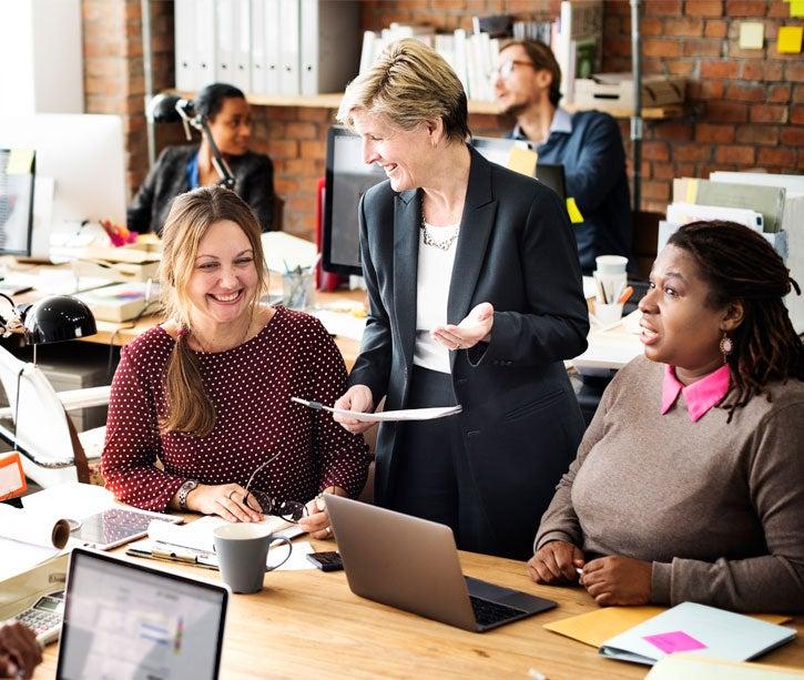 A group of intergenerational peers are in a huddle at work reading something on a laptop and smiling.
