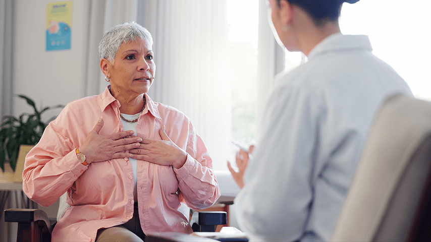 woman with short gray hair touching chest and talking to woman in white lab coat
