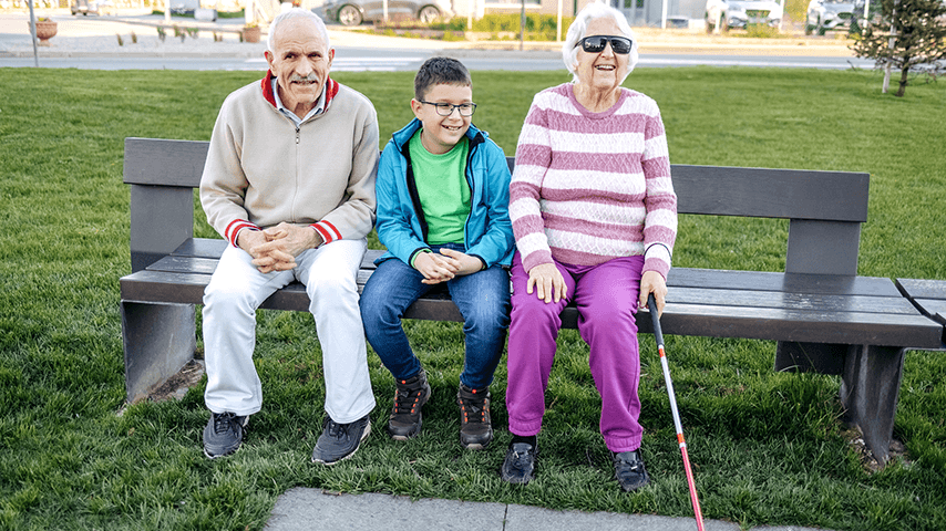 smiling grandmother wearing sunglasses and holding red-tipped cane seated next to grandson and grandfather on park bench