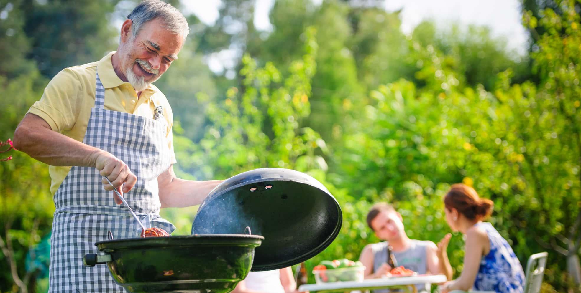 A senior Caucasian man is grilling outside while his family is enjoying a discussion on their patio.