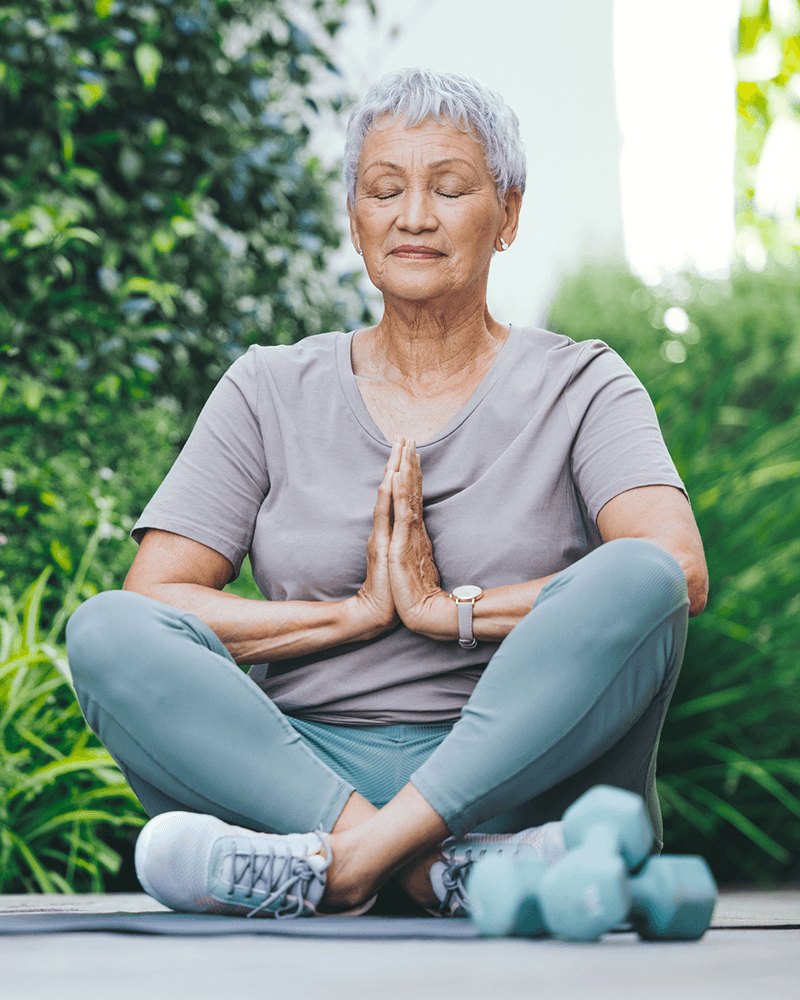An older woman with short gray hair sits cross-legged on the ground outdoors, meditating with her hands in a prayer position. She is wearing a light gray t-shirt, matching leggings, and athletic shoes. Her eyes are closed, and she appears calm and focused. In the foreground, a pair of teal dumbbells rests on the ground beside her. The background is filled with lush green foliage.