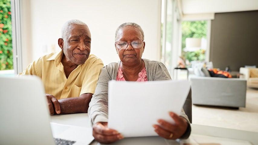 A man and a woman sit in front of a laptop computer sitting at a table. The women is holding a piece of paper and they are both looking at it.
