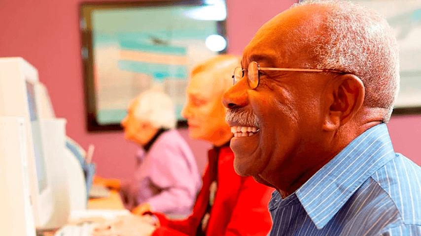 smiling older man at computer with two older women on computers behind him