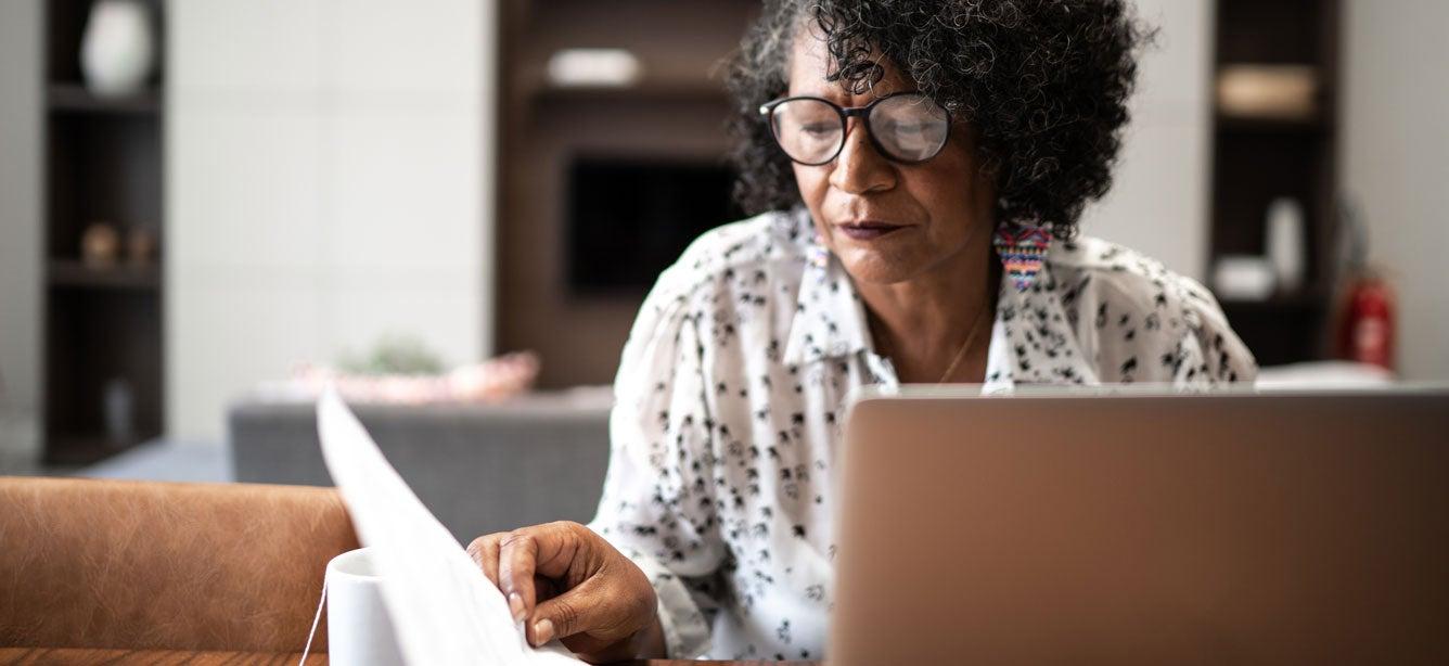 A Black senior woman is at her kitchen table, looking at her financials and working on her laptop.