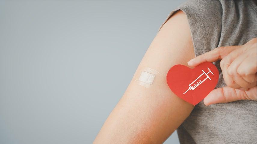 A woman's arm has a bandaid on it, where she's holding a red heart with a needle illustrated inside of it next to her bandaid, indicating that she received a flu shot to protect her health.