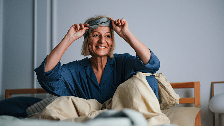 smiling middle-aged woman sitting up in bed removing eye mask