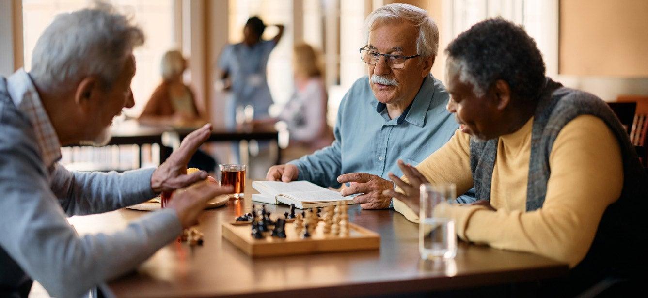 Three older men sitting at a table engaged in conversation, with a chessboard in front of them.