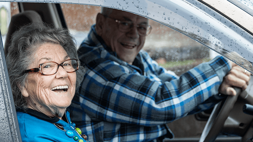 older adult man and woman smiling through the open window as they sit in their car on a rainy day