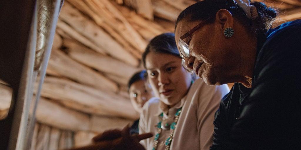 A senior Native American woman is showing a younger generation how to weave.