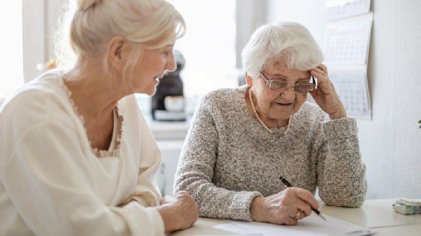 A caucasian senior woman is working on her find, end-of-life costs with her caregiver at a kitchen table in her home.