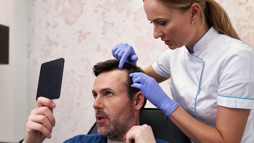 man holding and looking into hand mirror while woman in surgical gloves points at his receding hairline