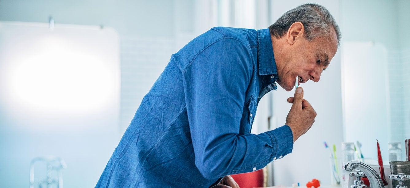 A middle-aged man with gray hair brushes his teeth while bending over a sink in a bright bathroom.